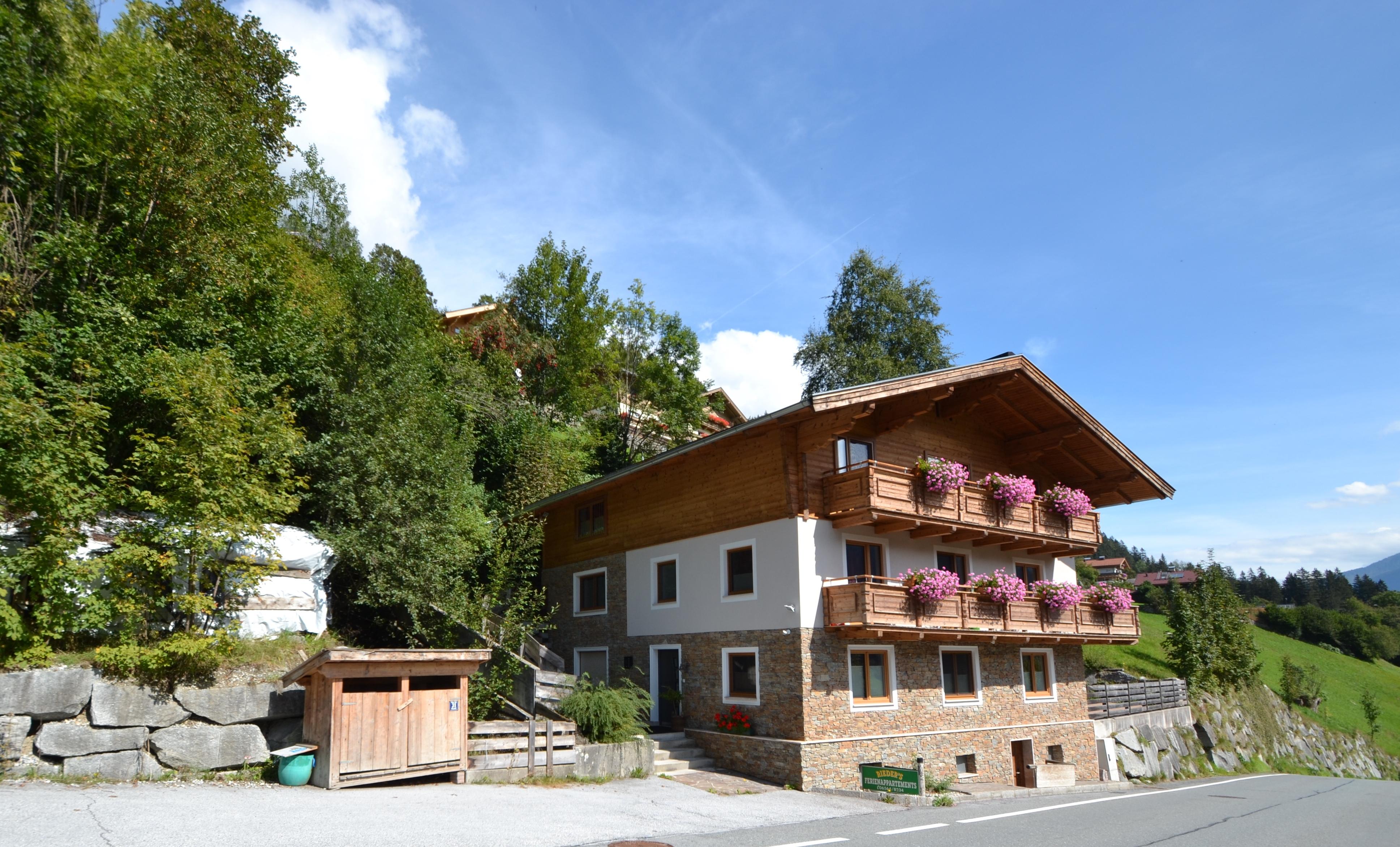 A traditional house in the mountains with a wooden upper floor and colorful flowers in the window. Surrounded by green forests and a clear blue sky.