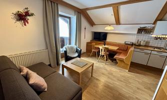 A cozy room with a sofa and a dining table. The wooden ceiling and large windows create a bright and inviting atmosphere.