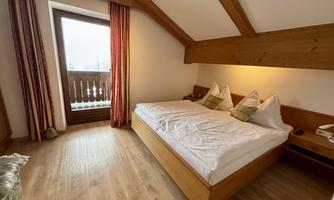 A cozy room with a double bed and wooden paneling. Through the window, a balcony is visible that offers a beautiful view.
