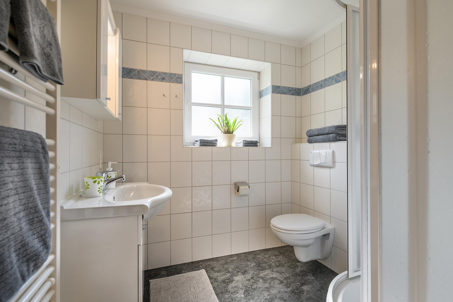 A modern bathroom with a bright tile wall and a window. It features a toilet, a sink, and a shower. Towels and bath towels, a hairdryer, and soap are provided. The Alpentherme Gastein, the Felsentherme Gastein, and the Gasteiner Heilstollen can be reached by car in a few minutes.