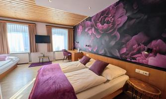 A modern room with a large bed and a purple flower wall. There is plenty of daylight coming through the windows and a cozy decor.