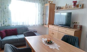 A cozy living room with a table, chairs, and a television. Large windows let in a lot of light.