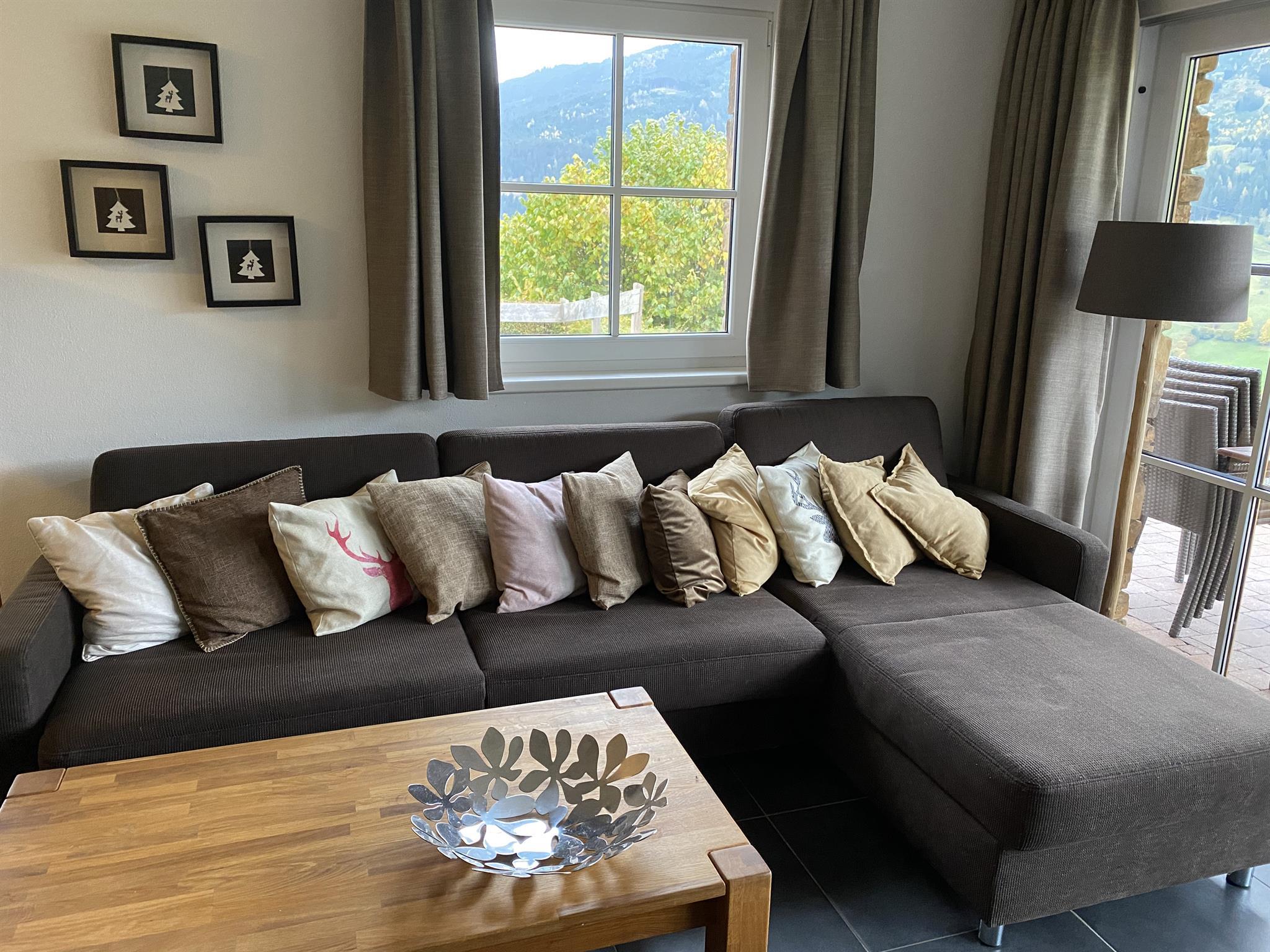A cozy living room with a brown sofa, decorated with colorful cushions. In the background, there are windows with a view of nature.