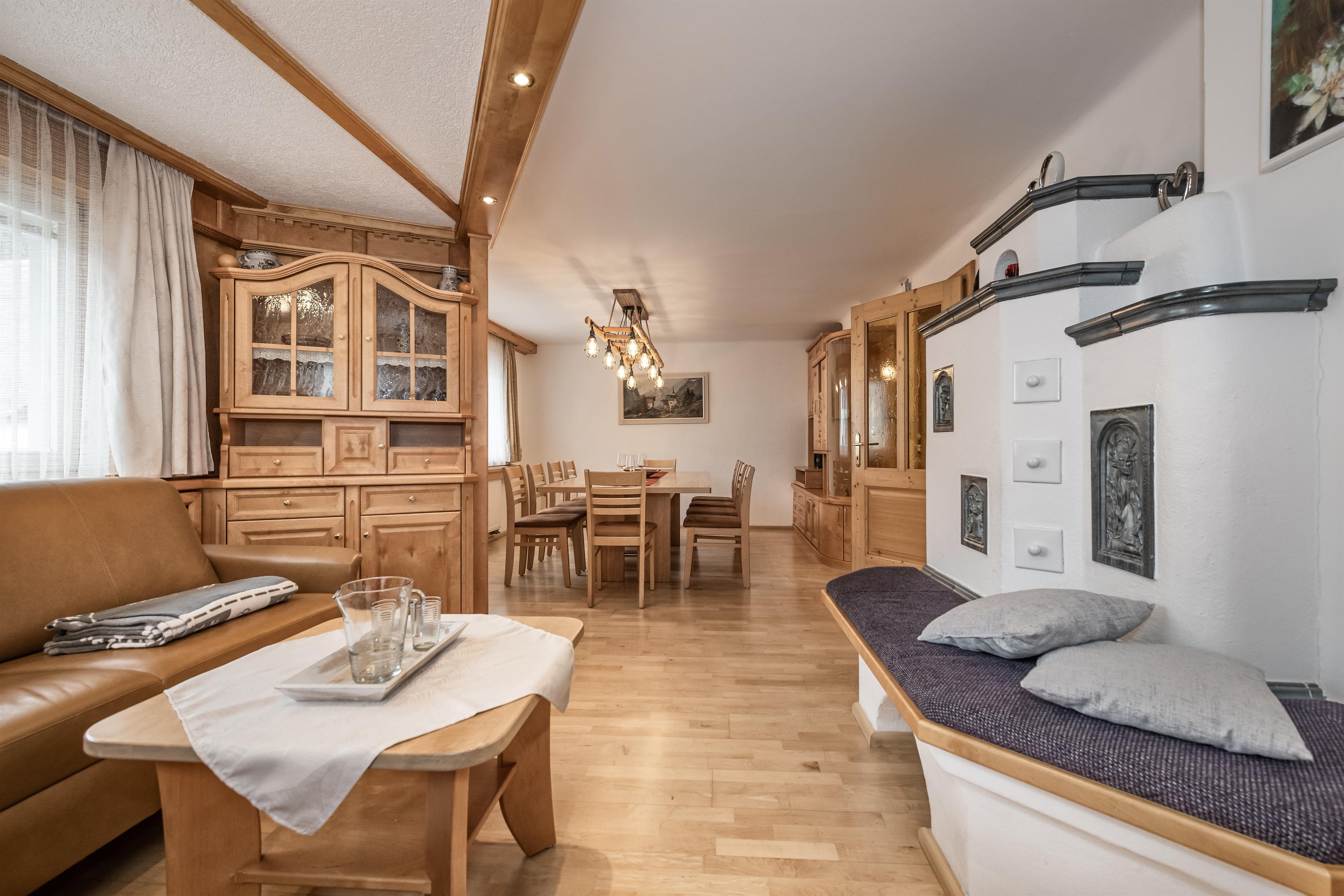 A cozy living area that was custom-made by the master carpenter with wooden furniture and offers an inviting atmosphere. The room features a dining table, seating area, tiled stove, TV, and stereo system, and is surrounded by windows and natural light with a view of the nearby ski slopes and mountains of Bad Hofgastein.