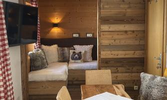 A cozy living space with wooden walls and a large table. On the left side, there is a comfortable seating area with cushions.