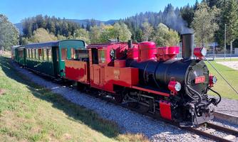An old steam locomotive in red with a green carriage is standing on the tracks. It is surrounded by a beautiful landscape of trees and hills.