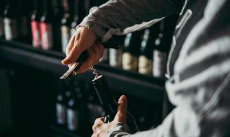 A person opens a bottle of wine with a corkscrew. Many wine bottles are visible in the background.
