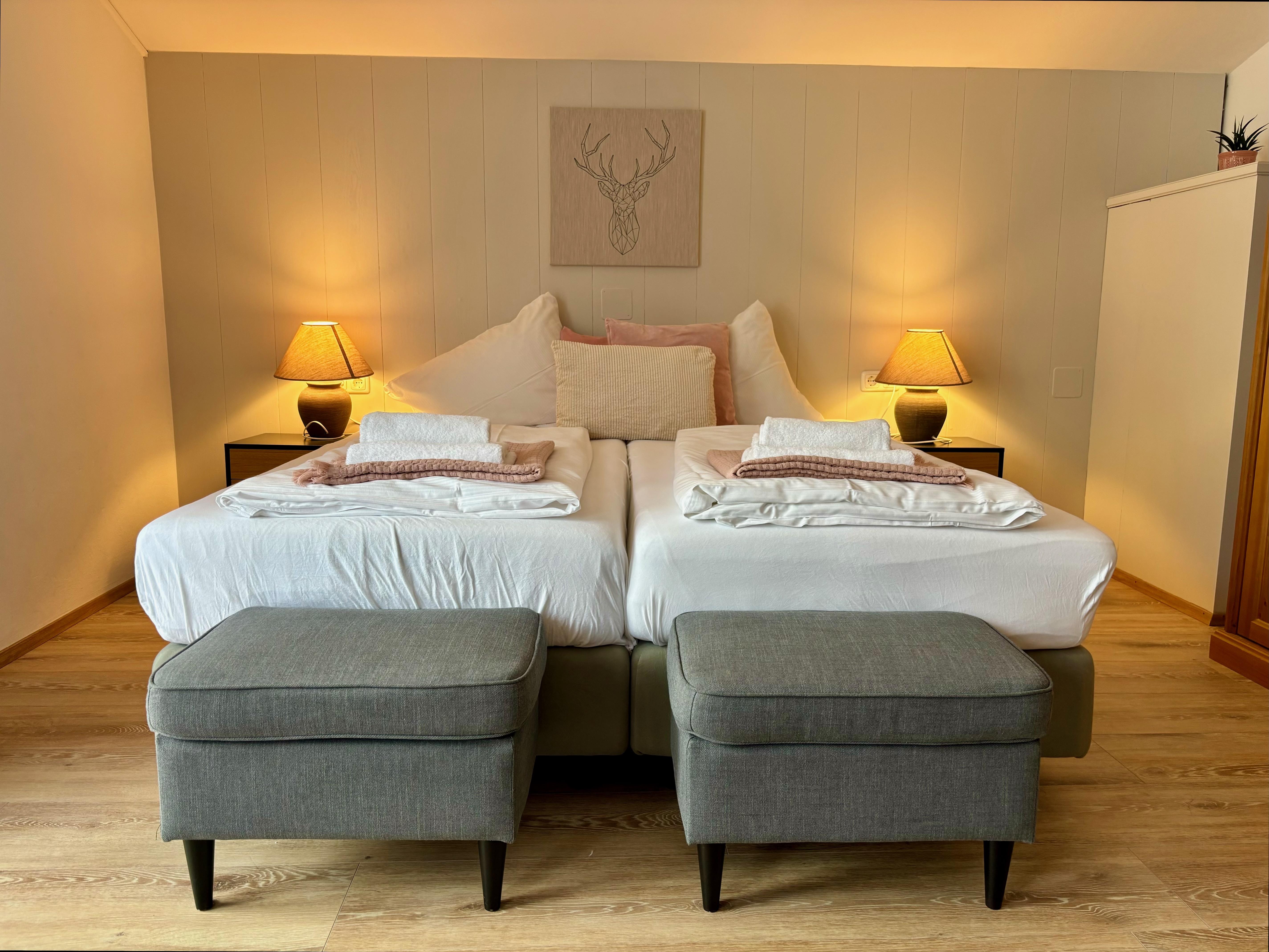A cozy bedroom with a large double bed and two gray stools. The warm lamp lamps create an inviting atmosphere.