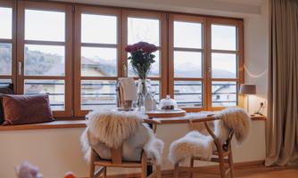 A cozy dining area with a table and chairs covered in fur. Large windows provide a view of the mountains and let in plenty of light.