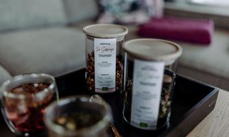 Two glass jugs with different types of tea are on a tray. In the background, there are glasses with drinks and a cozy living area visible.