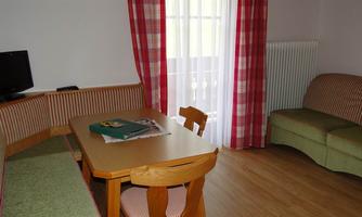 A cozy living space with a dining table, a couch, and a television. Large windows let in plenty of light, and the curtains give the room a friendly look.