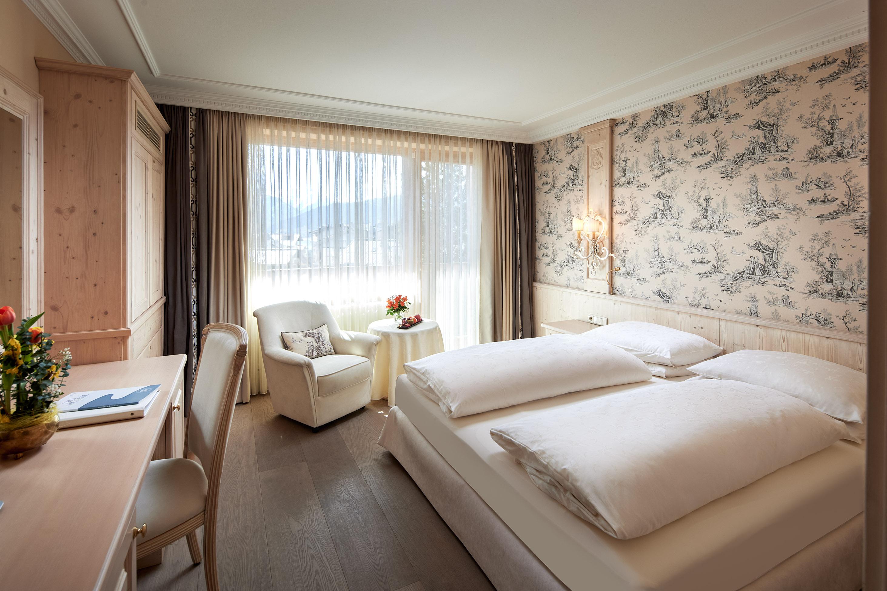 A cozy hotel room with a large double bed and a bright window. There is a comfortable armchair and a small table decoration.