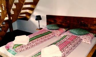 A cozy bedroom with a large wooden bed and colorful blankets. A wooden staircase leads to the upper floor, and a small nightstand stands next to the bed.