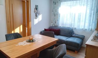 A cozy living space with a wooden dining table and a gray sofa. Light streams through a large window with white curtains.