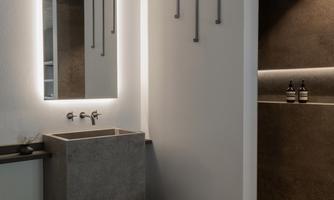 A modern bathroom with a minimalist sink arrangement and a large mirror. The walls are made of dark concrete, and there is a small wooden stool.