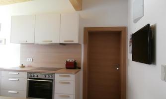 A modern kitchen with white cabinets and a wooden ceiling. In the background, a door and a television on the wall are visible.