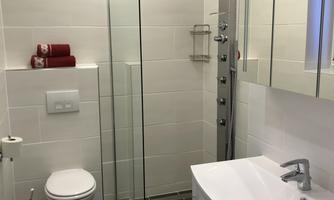 A modern bathroom with a glass shower enclosure and an elegant vanity unit. The walls are tiled in light colors and the floor has a gray tile pattern.