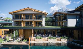 A modern building with a wood facade and large windows. In the foreground, there is a tranquil water area with seating.