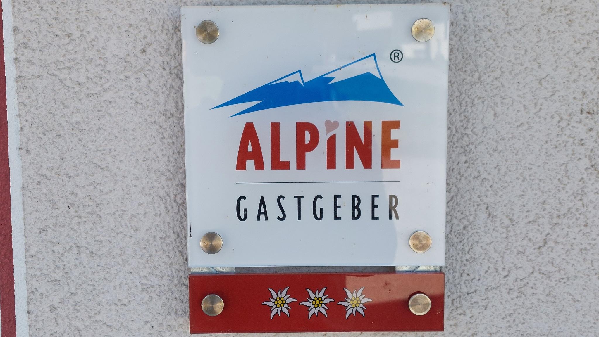 A sign with the inscription "ALPINE HOST". It features a mountain silhouette and floral patterns below.