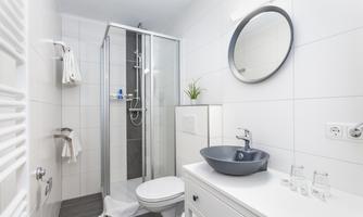 A modern bathroom with a shower and a sink. The walls are tiled in white and there is a round mirror.