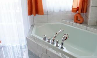 A modern bathroom with a corner bathtub and orange curtains. The floor is tiled and there is a rug in soft colors.