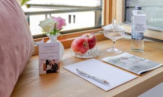 A cozy window seat with a vase of flowers, apples, water, and writing materials. There is also a brochure and a notepad on the table.