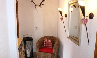 A cozy entrance area with a woven chair and a red cushion. On the wall hang stag antlers and decorative wall lamps.
