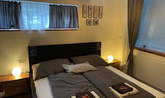 A cozy bedroom with a double bed and soft lighting. The walls are light-colored and there are windows with curtains.