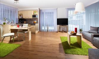 A modern living room with light wooden floors and large windows. The room is furnished with a dining table, a sofa, and green carpets.