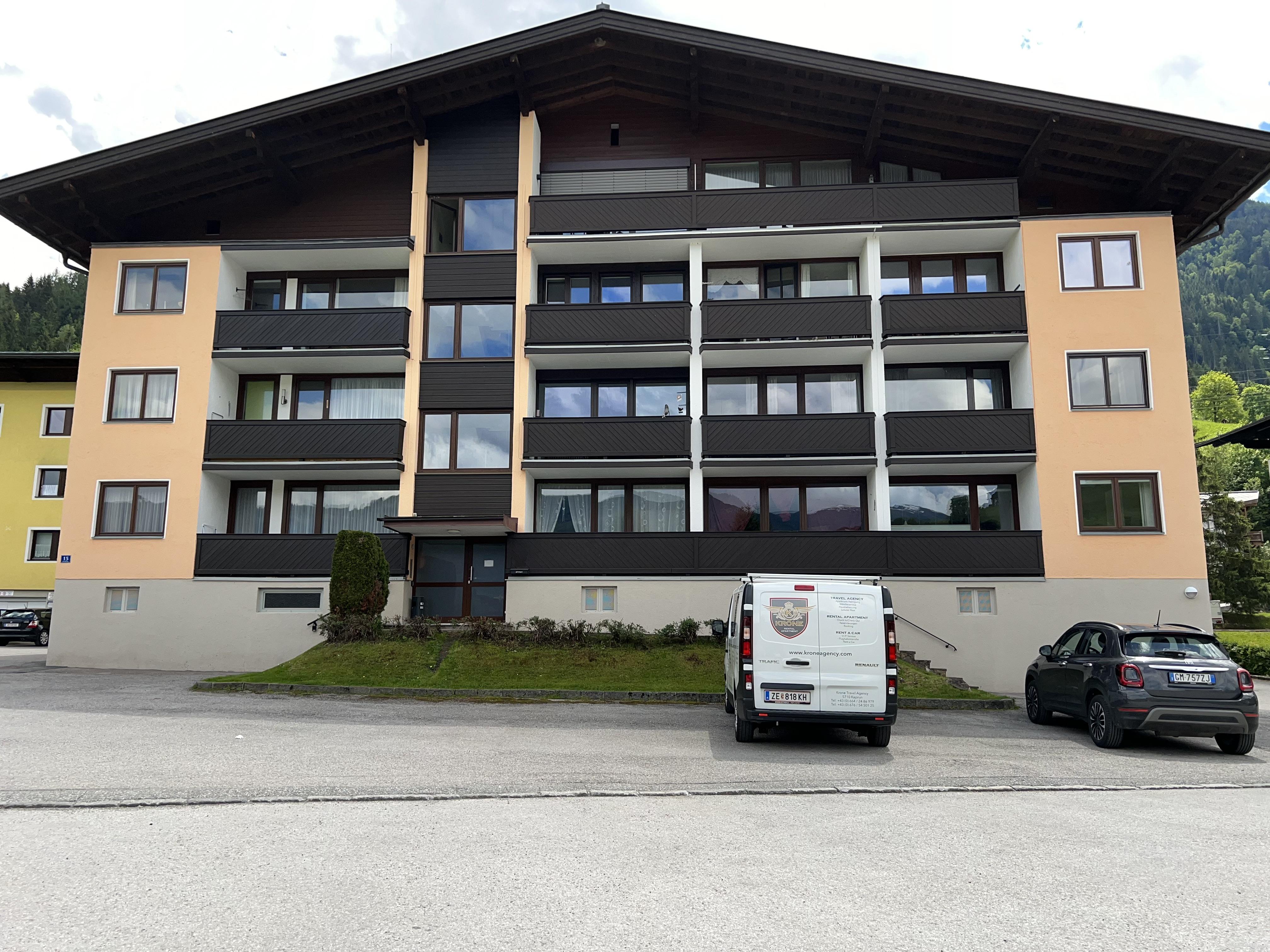 A modern residential building with multiple floors and balconies. In front of the building, there is a delivery van and a car in the parking lot.