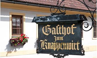 A traditional inn sign with the inscription "Galthof zum Knappenwirt". In the background, windows with flower boxes can be seen.