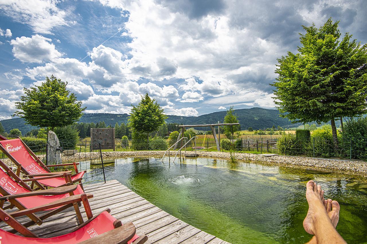 A tranquil view of a pond with green water and cozy loungers. The landscape is surrounded by trees and mountains under a striking sky with clouds.