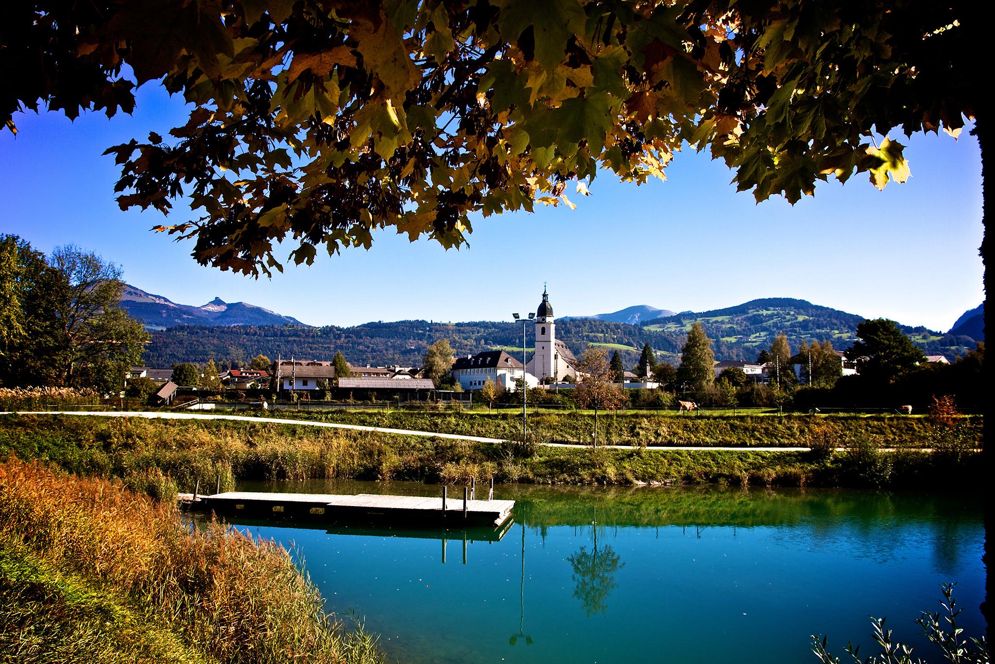 A serene lake surrounded by colorful trees and mountains. In the background, a small church can be seen.