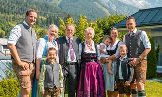 A cheerful family stands outdoors in traditional clothing. In the background, green mountains and a clear blue sky are visible.