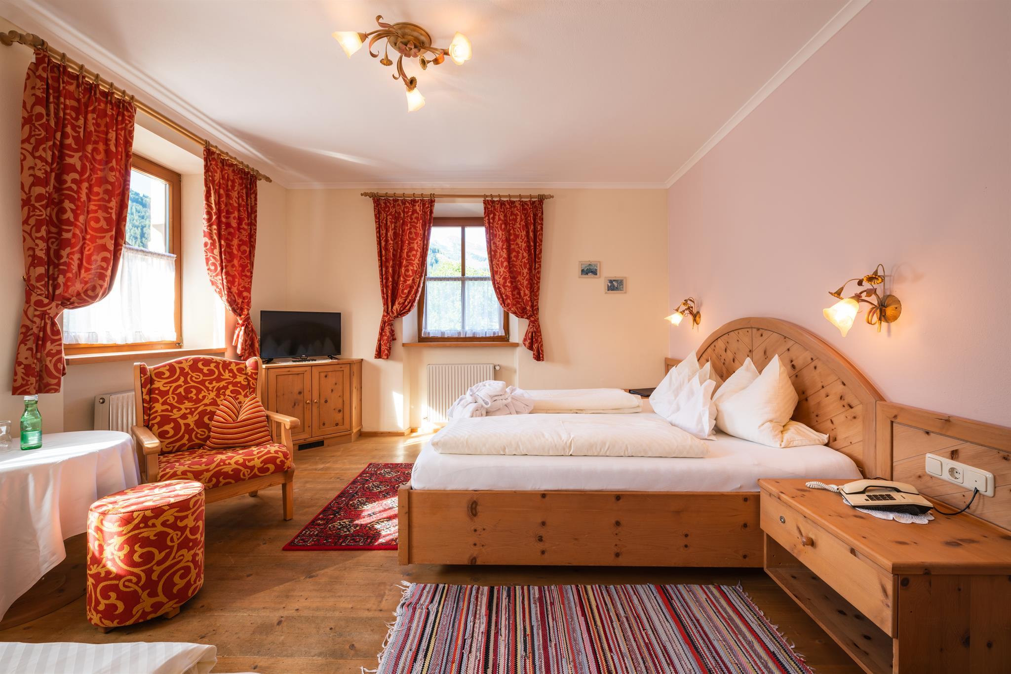 A cozy room with two beds and a large window. It is furnished with wooden furniture and colorful curtains.