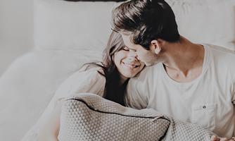A couple sits on a bed and smiles at each other. They appear happy and intimate as they hug a pillow.