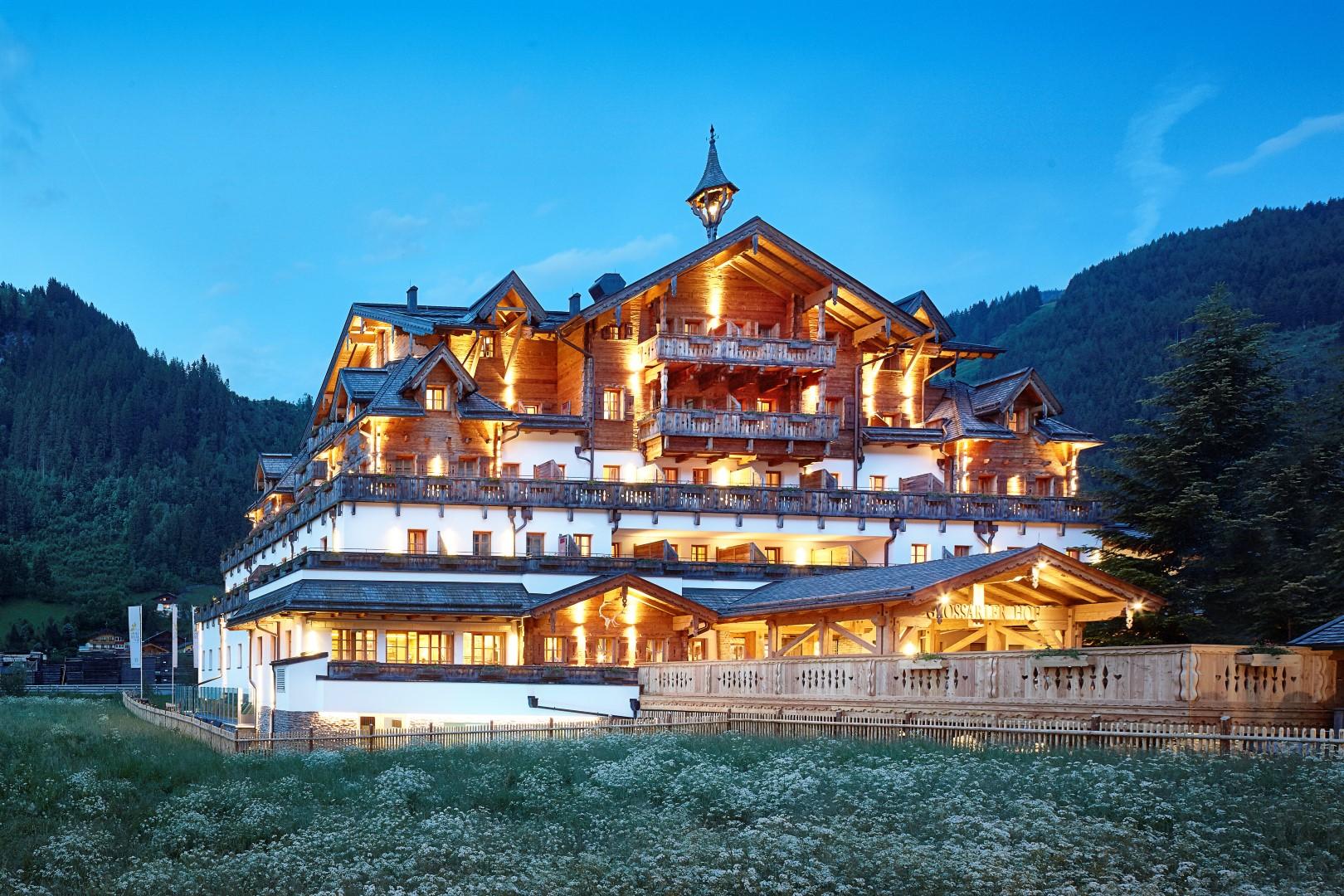 A beautiful mountain hotel with many balconies and warm lighting. Surrounded by green meadows and mountains in the background.