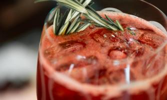A red cocktail with ice cubes and a sprig of rosemary. The drink is served in an elegant glass.
