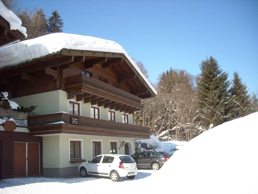 A cozy wooden house in the snow with a large roof. Surrounded by snow-covered trees and clear sky.
