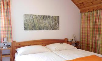 A cozy bedroom with a double bed and wooden ceiling. The windows are adorned with checkered curtains, and a picture of grasses hangs on the wall.
