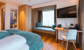A modern bedroom with a comfortable bed and a cozy window seat. There is a desk, a television, and warm wooden surfaces.