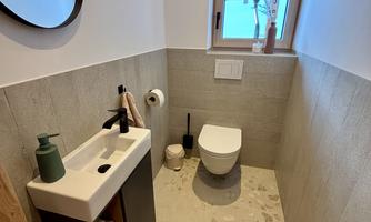 A modern bathroom with a sink, a toilet area, and a small window. The walls are kept light, and the floor has a light texture.