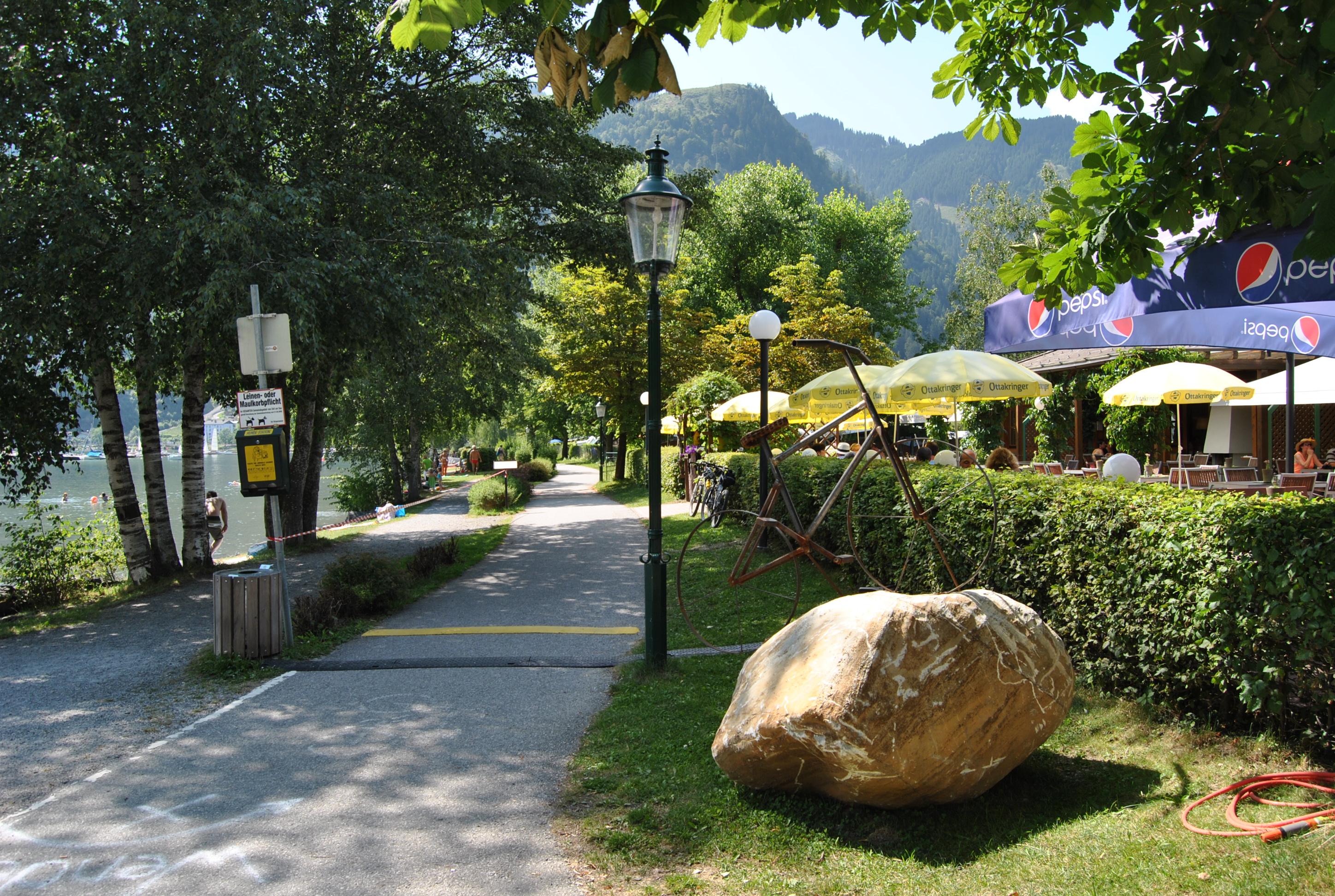 A beautiful path along a lake with trees and seating areas. In the background, a restaurant and mountains can be seen.