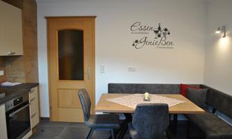 A modern kitchen with a dining table and seating benches. Decorative lettering hangs on the wall.