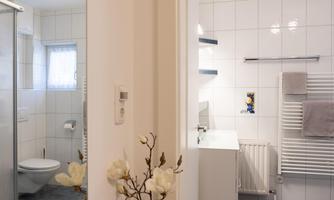 A modern bathroom with white tiles and high-quality fixtures. On the left side, there is a shower, and on the right, a sink with towels.