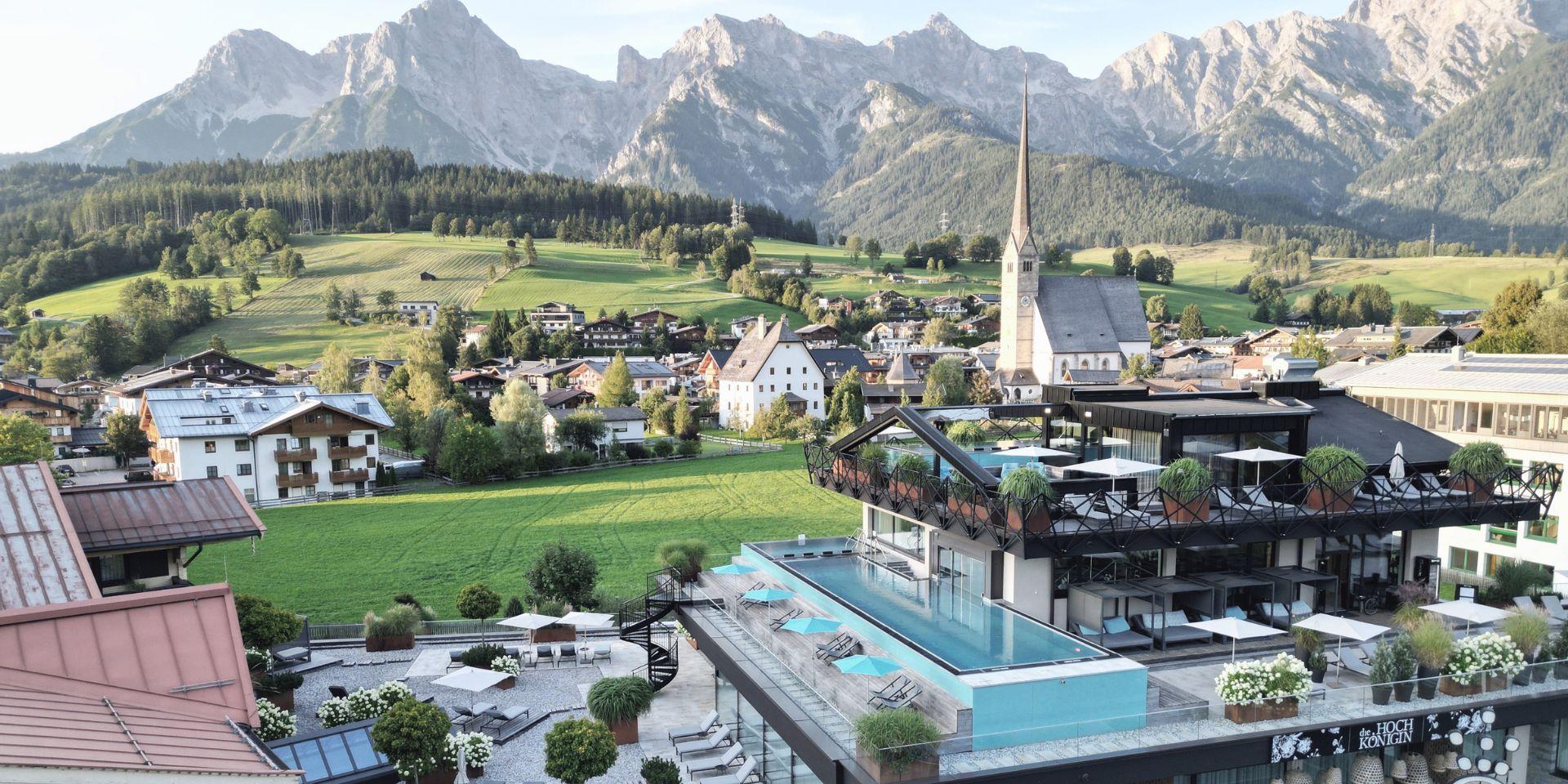 A picturesque landscape with mountains in the background and a quiet village in the foreground. At the edge of the village, there is a modern building with a pool.