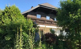 A large, rustic house surrounded by lush greenery and various plants. The sky is clear and radiant blue.