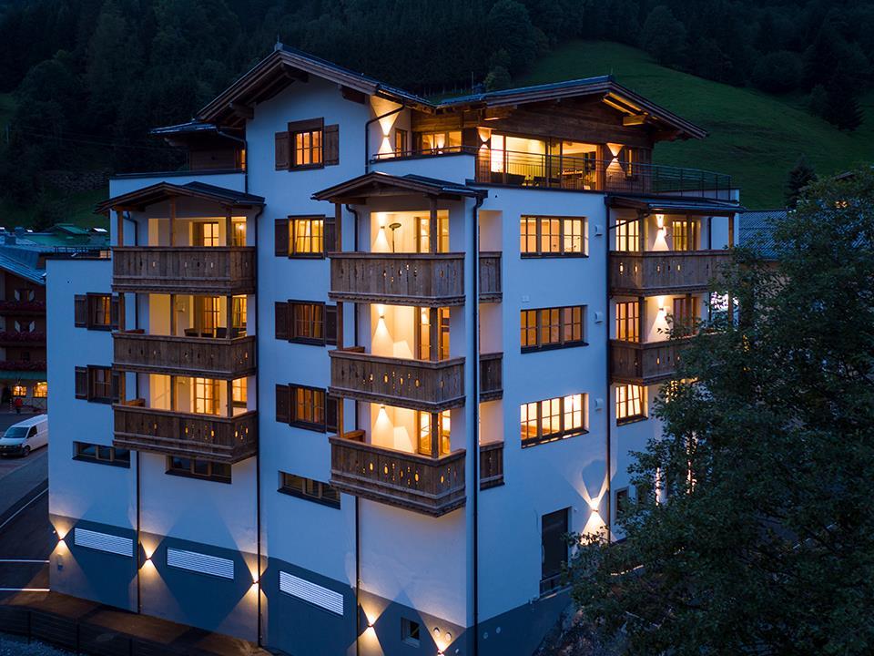 A modern building with wooden cladding and many windows, illuminated in the evening hours. It is located in a mountainous setting.
