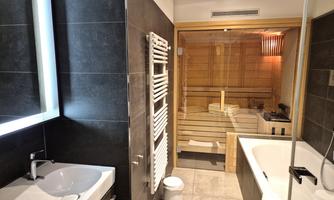 A modern bathroom with a bathtub and a shower. In the background, there is a wooden sauna.