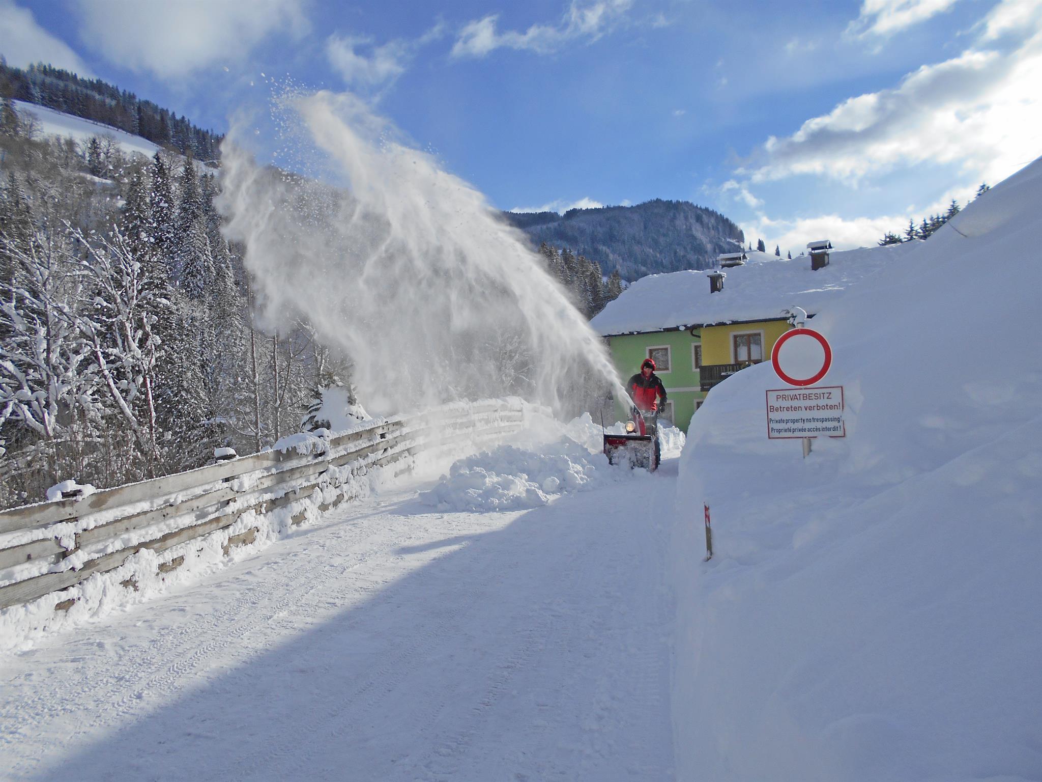A snowplow clears a snowy path in the mountains. The scene shows fresh snow and a clear blue sky.
